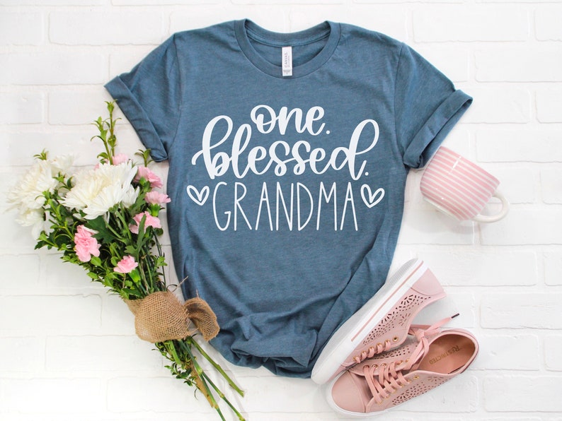 May include: A blue t-shirt with the text "one blessed grandma" in white lettering with two white hearts on either side of the word "grandma".