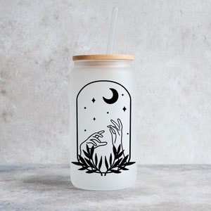 May include: Frosted glass can-shaped tumbler with a bamboo lid and a clear straw. The tumbler features a black graphic of hands reaching towards a crescent moon and stars within an arched frame, with a leafy design at the base.