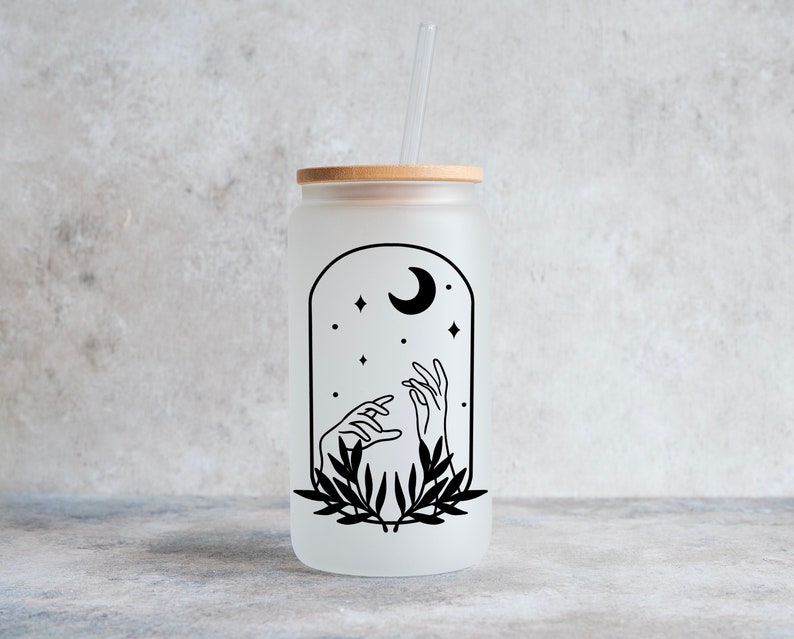 May include: Frosted glass can-shaped tumbler with a bamboo lid and a clear straw. The tumbler features a black graphic of hands reaching towards a crescent moon and stars within an arched frame, with a leafy design at the base.