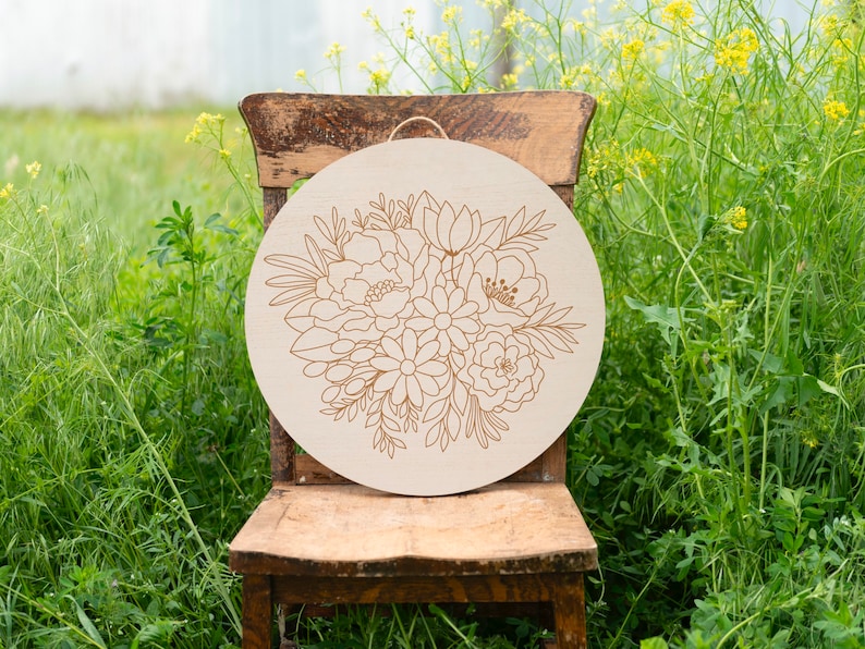 May include: A round wooden sign with a floral design. The design features a bouquet of flowers in a light brown color. The sign is hanging on a wooden chair.