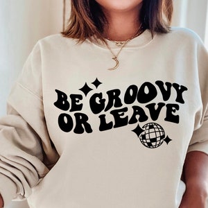 May include: A beige sweatshirt with the text "Be groovy or leave" in black, retro-style font. A black disco ball graphic is below the text.