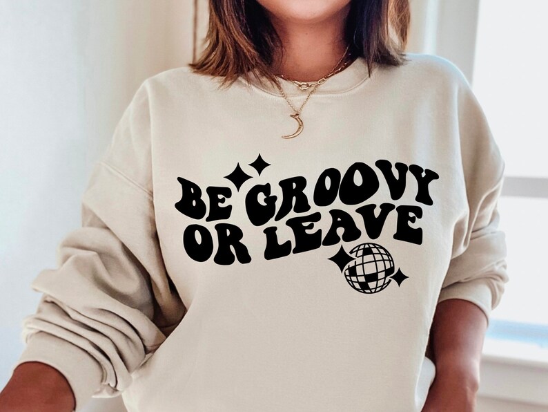 May include: A beige sweatshirt with the text "Be groovy or leave" in black, retro-style font. A black disco ball graphic is below the text.