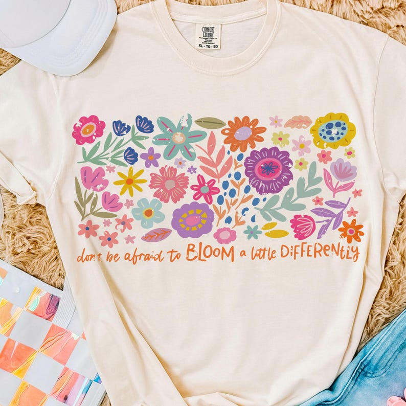 May include: A cream-colored t-shirt with a colorful floral design and the text "don't be afraid to BLOOM a little DIFFERENTLY".