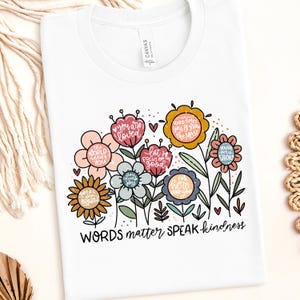 May include: White t-shirt with a floral design and the text "WORDS matter SPEAK kindness". The flowers are in various colors and have positive messages written on them, such as "you are loved", "the world needs you", and "you deserve joy".