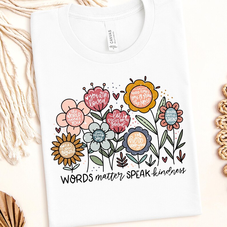 May include: White t-shirt with a floral design and the text "WORDS matter SPEAK kindness". The flowers are in various colors and have positive messages written on them, such as "you are loved", "the world needs you", and "you deserve joy".