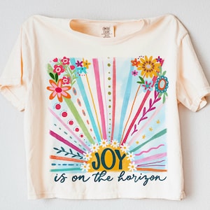 May include: A light yellow t-shirt with a colorful graphic of a sunrise with flowers and the text "JOY is on the horizon".