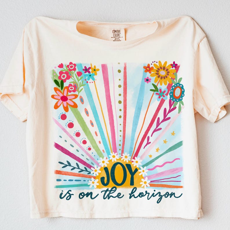 May include: A light yellow t-shirt with a colorful graphic of a sunrise with flowers and the text "JOY is on the horizon".