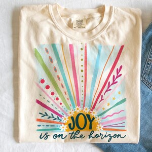 May include: A cream colored t-shirt with a colorful sunrise graphic and the text "JOY is on the horizon".