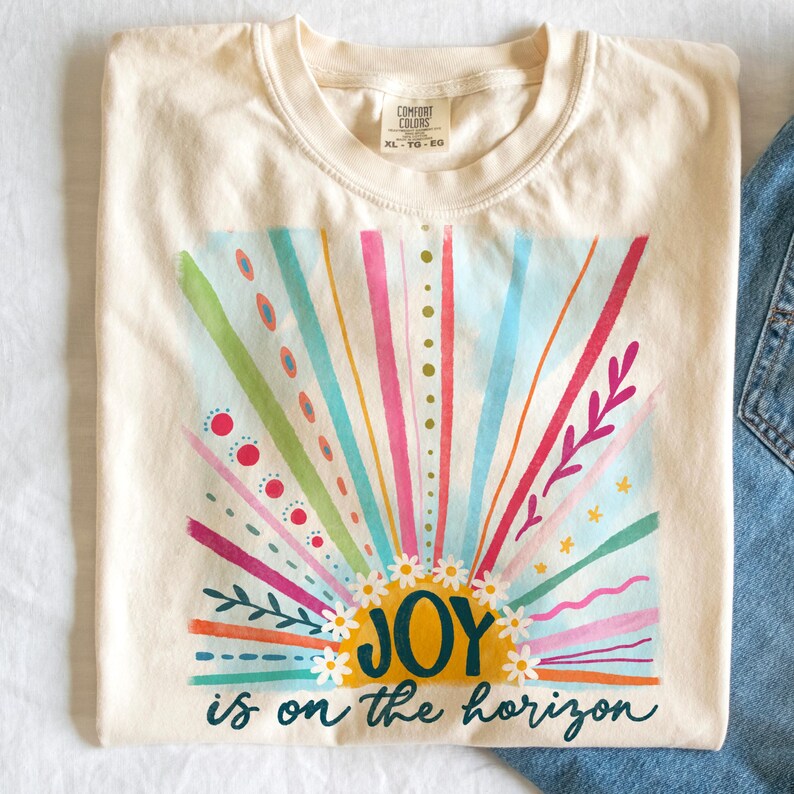 May include: A cream colored t-shirt with a colorful sunrise graphic and the text "JOY is on the horizon".