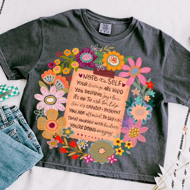 May include: Dark gray t-shirt with a floral design surrounding a note that reads "NOTE to SELF" and positive affirmations. The shirt is paired with blue jeans and a checkered accessory.