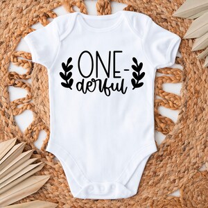 Op de afbeelding: Witte baby romper met zwarte tekst die "ONE-derful" luidt met bladeren aan beide kanten van de tekst.