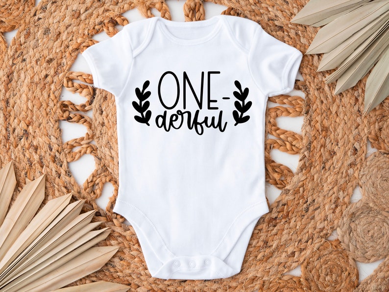 Op de afbeelding: Witte baby romper met zwarte tekst die "ONE-derful" luidt met bladeren aan beide kanten van de tekst.