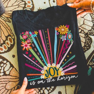 May include: Black t-shirt with a colorful graphic of a sun with the word "JOY" inside and the text "is on the horizon" below. The graphic features a rainbow of colors and floral accents.