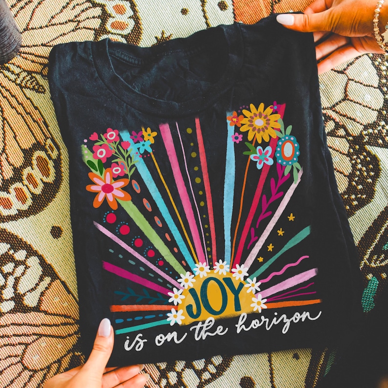 May include: Black t-shirt with a colorful graphic of a sun with the word "JOY" inside and the text "is on the horizon" below. The graphic features a rainbow of colors and floral accents.