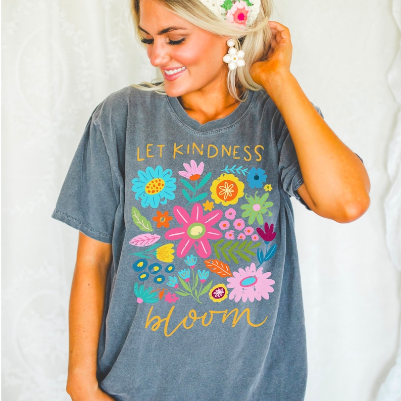 Peut inclure: T-shirt gris avec un motif floral color&eacute; et le texte "Let Kindness Bloom" en lettres dor&eacute;es.
