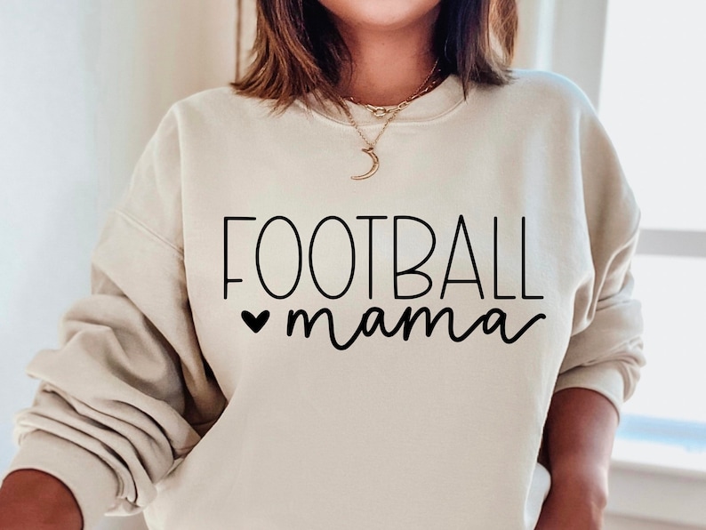 May include: A cream-colored sweatshirt with the text "FOOTBALL mama" in black lettering. A small heart is in front of the word "mama".