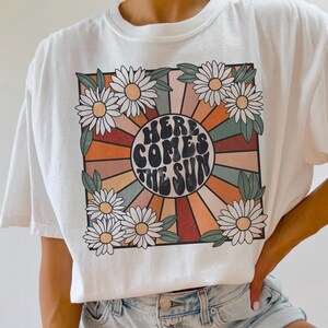 May include: White t-shirt with a retro-style graphic featuring a sunburst design with the text "Here Comes The Sun" in a circle surrounded by daisies.