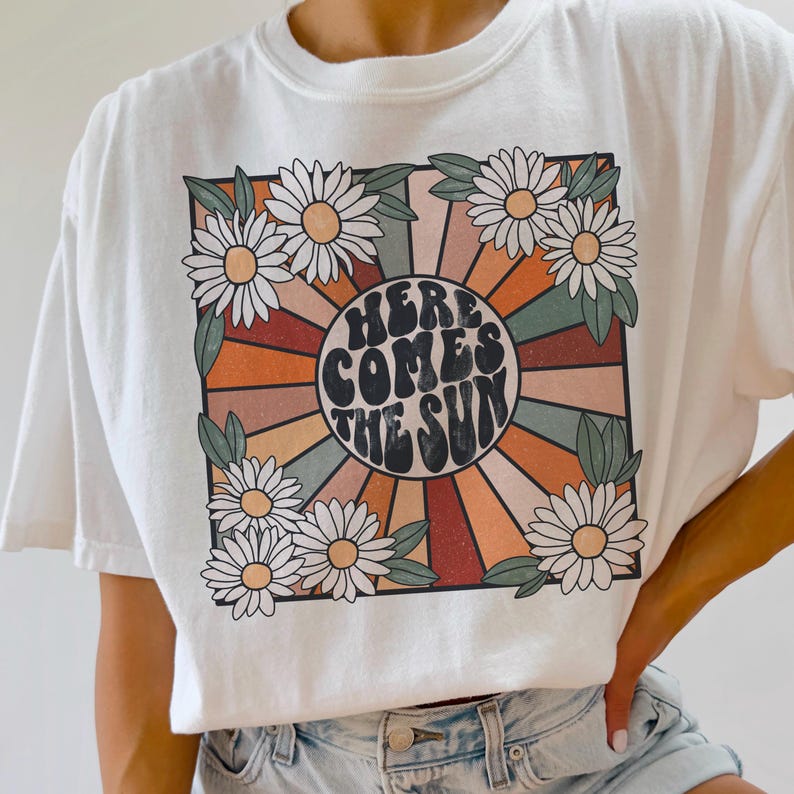 May include: White t-shirt with a retro-style graphic featuring a sunburst design with the text "Here Comes The Sun" in a circle surrounded by daisies.
