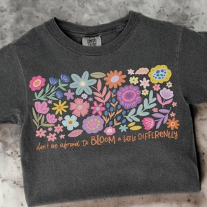 May include: A dark grey t-shirt with a colorful floral design and the text "don't be afraid to BLOOM a little DIFFERENTLY".