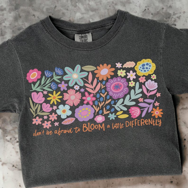 May include: A dark grey t-shirt with a colorful floral design and the text "don't be afraid to BLOOM a little DIFFERENTLY".