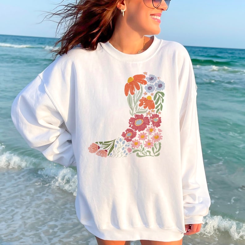 May include: White sweatshirt with a floral design in the shape of a cowboy boot. The flowers are in shades of pink, red, orange, yellow, and blue.