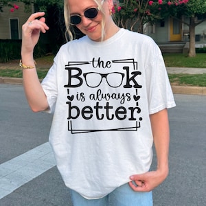 May include: White t-shirt with black text that reads "the Book is always better." The text is framed with a black border and includes a pair of glasses as part of the word "Book".