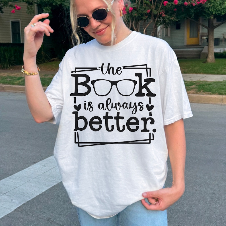 May include: White t-shirt with black text that reads "the Book is always better." The text is framed with a black border and includes a pair of glasses as part of the word "Book".