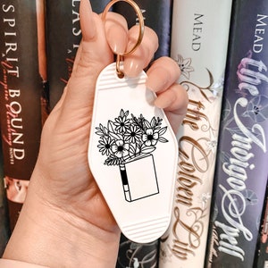 May include: White keychain shaped like a motel key with a black and white illustration of a book with flowers coming out of the top. The text "The Bookish Life" is printed on the keychain.