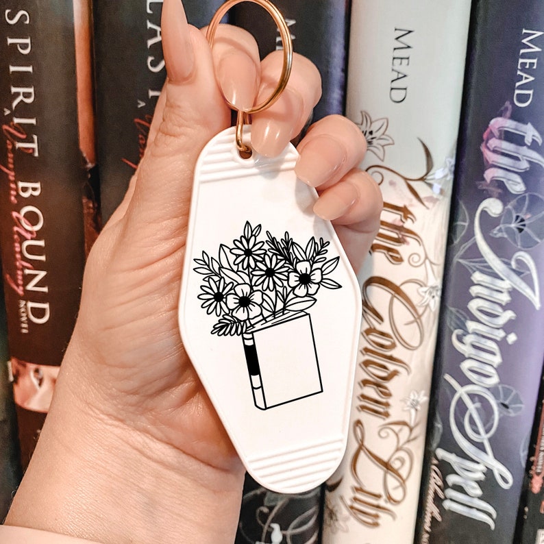 May include: White keychain shaped like a motel key with a black and white illustration of a book with flowers coming out of the top. The text "The Bookish Life" is printed on the keychain.