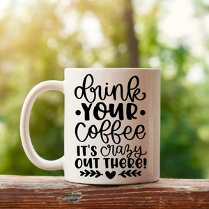 K&ouml;nnte beinhalten: Wei&szlig;e Keramik-Kaffeetasse mit schwarzem Text, der "drink your coffee it's crazy out there!" lautet, mit einem kleinen Herz und einem Blattdesign am Boden.