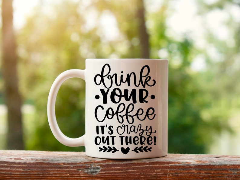 K&ouml;nnte beinhalten: Wei&szlig;e Keramik-Kaffeetasse mit schwarzem Text, der "drink your coffee it's crazy out there!" lautet, mit einem kleinen Herz und einem Blattdesign am Boden.