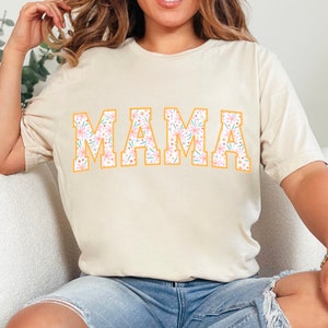 Op de afbeelding: Een beige T-shirt met een bloemenprintontwerp dat "MAMA" spelt in grote, gedurfde letters met een gele omtrek.