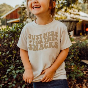 K&ouml;nnte beinhalten: Ein beiges T-Shirt mit dem Text "JUST HERE FOR THE SNACKS" in brauner Retro-Schrift. Das Shirt hat einen Rundhalsausschnitt und kurze &Auml;rmel. Das Bild zeigt ein Kind, das das Shirt tr&auml;gt.