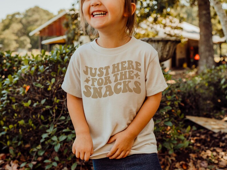 K&ouml;nnte beinhalten: Ein beiges T-Shirt mit dem Text "JUST HERE FOR THE SNACKS" in brauner Retro-Schrift. Das Shirt hat einen Rundhalsausschnitt und kurze &Auml;rmel. Das Bild zeigt ein Kind, das das Shirt tr&auml;gt.
