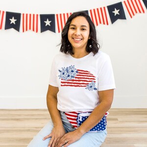 K&ouml;nnte beinhalten: Ein wei&szlig;es T-Shirt mit einem rot-wei&szlig;-blauen amerikanischen Flaggenmuster mit blauen Blumen. Das Shirt wird von einer Person mit braunen Haaren und einer wei&szlig;en Bauchtasche mit einem rot-wei&szlig;-blauen amerikanischen Flaggenmuster getragen.