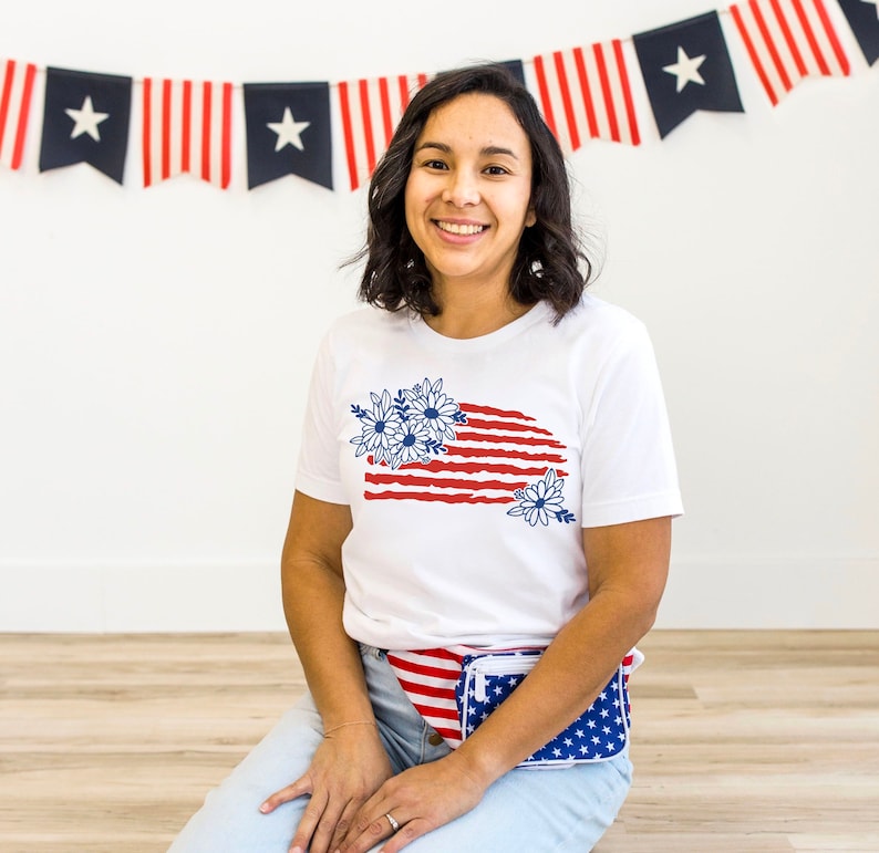 K&ouml;nnte beinhalten: Ein wei&szlig;es T-Shirt mit einem rot-wei&szlig;-blauen amerikanischen Flaggenmuster mit blauen Blumen. Das Shirt wird von einer Person mit braunen Haaren und einer wei&szlig;en Bauchtasche mit einem rot-wei&szlig;-blauen amerikanischen Flaggenmuster getragen.