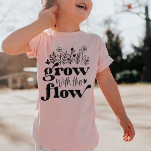K&ouml;nnte beinhalten: Hellrosa T-Shirt mit dem schwarzen Schriftzug "grow with the flow". &Uuml;ber dem Text befindet sich ein Blumendesign. Das T-Shirt wird von einem Kind getragen.
