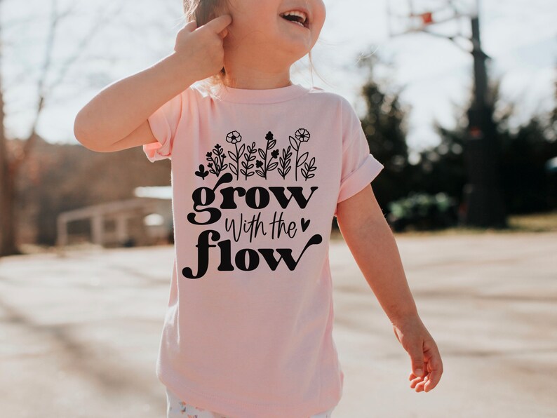 K&ouml;nnte beinhalten: Hellrosa T-Shirt mit dem schwarzen Schriftzug "grow with the flow". &Uuml;ber dem Text befindet sich ein Blumendesign. Das T-Shirt wird von einem Kind getragen.