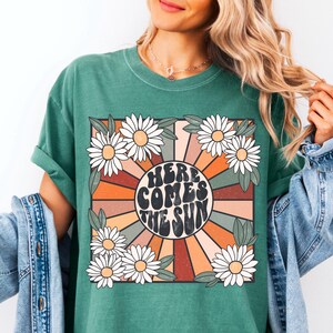 May include: Green t-shirt with a retro-style graphic featuring a sunburst design with the text "Here Comes The Sun" in a groovy font. The graphic is surrounded by white daisies.