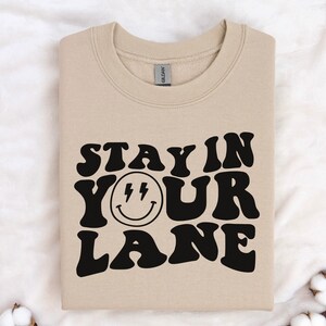 May include: A beige sweatshirt with the text "Stay in your lane" in black, stylized font. A black smiley face with lightning bolts for eyes is centered between the words "your" and "lane".