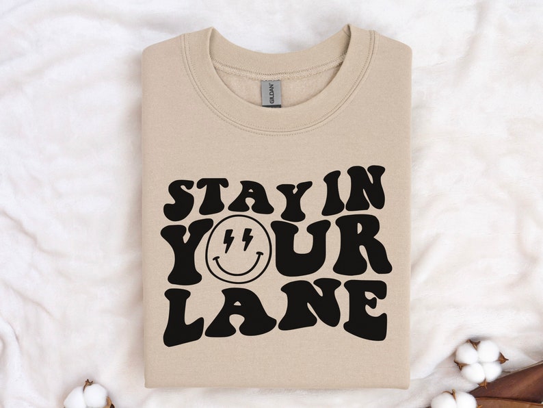 May include: A beige sweatshirt with the text "Stay in your lane" in black, stylized font. A black smiley face with lightning bolts for eyes is centered between the words "your" and "lane".
