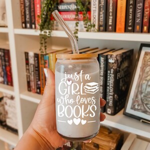 May include: A clear glass tumbler with a wooden lid and a straw. The tumbler has a white vinyl decal that says "just a girl who loves books" with a small heart above the word "girl" and three hearts below the word "books".