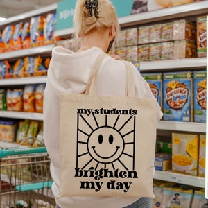 May include: A white canvas tote bag with black text that reads "my students brighten my day" and a black and white illustration of a smiling sun.