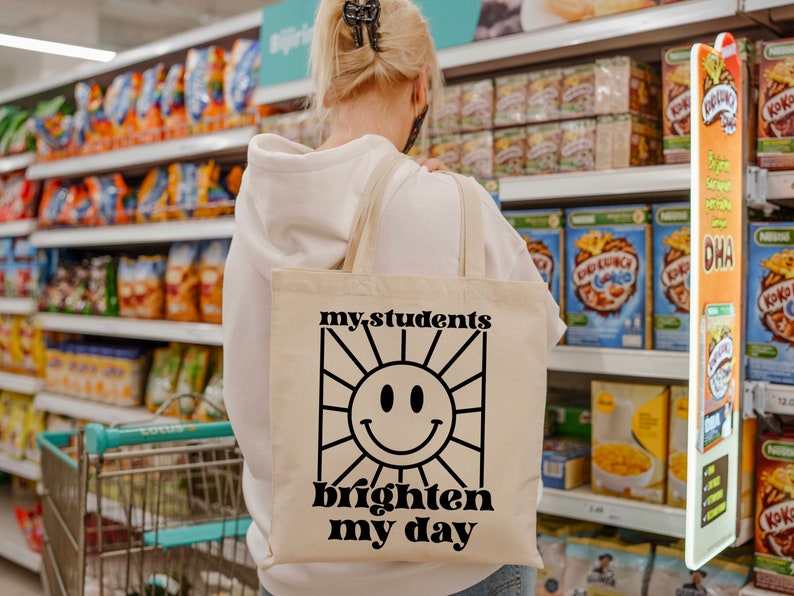 May include: A white canvas tote bag with black text that reads "my students brighten my day" and a black and white illustration of a smiling sun.