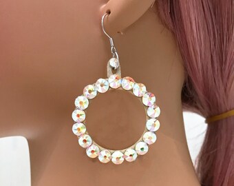May include: A pair of large, round, cream-coloured earrings with a silver hook. The earrings are adorned with a circle of iridescent rhinestones.