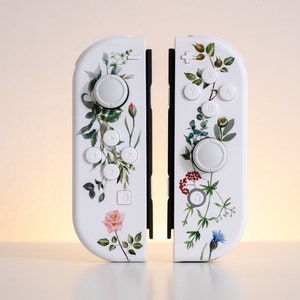 May include: A pair of white Nintendo Switch Joy-Con controllers with a floral design. The controllers have a white background with green leaves, pink and blue flowers, and other floral details.