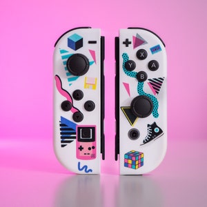 May include: A pair of white Nintendo Switch Joy-Con controllers with a retro 80s themed design. The controllers feature a variety of colorful geometric shapes, including a Rubik's Cube, a sneaker, and a pixelated video game character. The controllers are separated, showing the left and right sides.