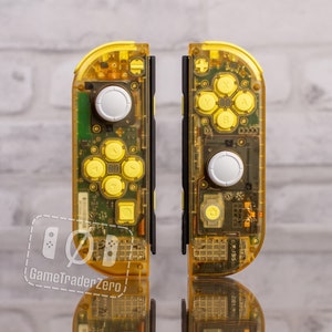 May include: Two transparent yellow Nintendo Switch Joy-Con controllers with white buttons. The controllers are on a white brick wall background.