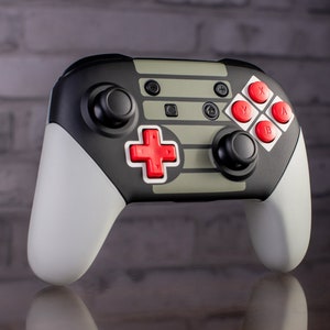 May include: A black and gray video game controller with red buttons. The controller has a gray and black design with the letters "A", "B", "X", and "Y" on the buttons. The controller is designed to look like a classic Nintendo Entertainment System controller.