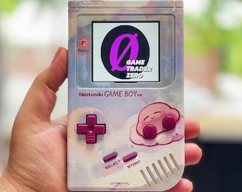 Puede incluir: Una Game Boy Color rosa y blanca con un diseño personalizado que presenta una "O" rosa con el texto "GAME TRADER ZERO" y un personaje rosa de Kirby. Los botones son morados.